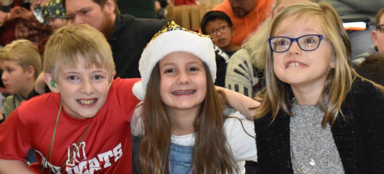 three children facing the camera smiling, middle child has a santa hat on