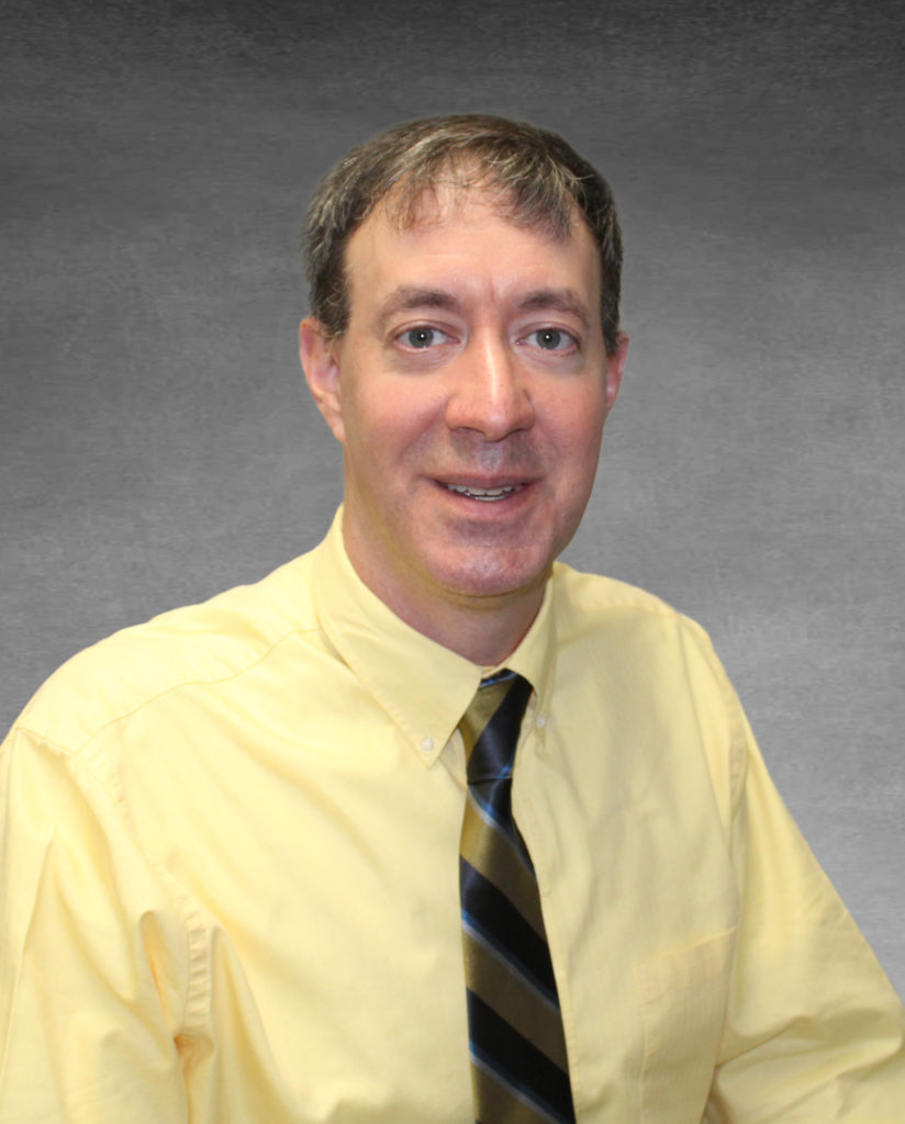 Professional headshot of Mike smiling. He is wearing a yellow button up shirt and a gold, black, silver, and blue striped tie