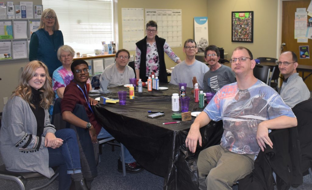 Photo of Tuesday Group members seated around a table and smiling for the camera. There are paints scattered around the table.