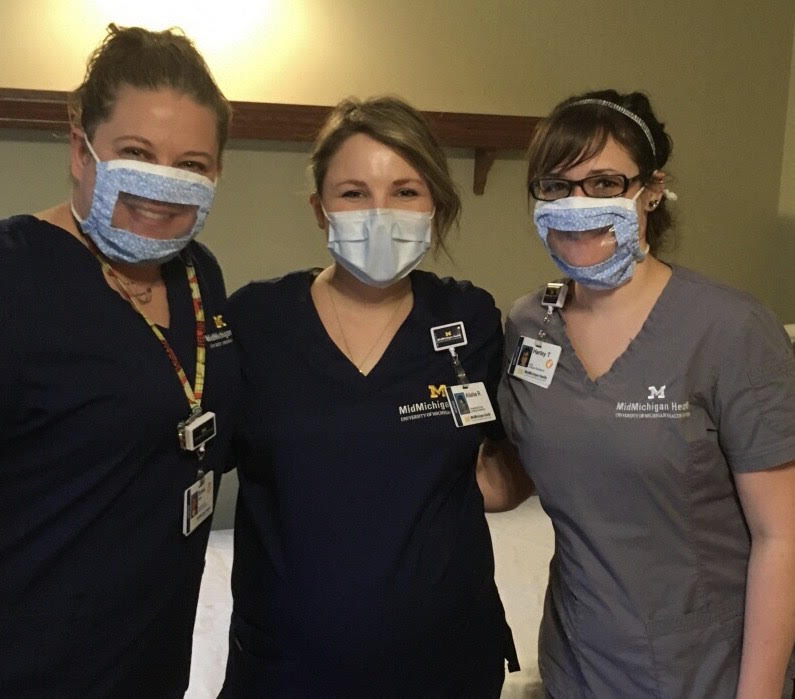 Three nurses standing together. The two on the end have face masks with clear mouth window. The one in the middle has a normal mask on.