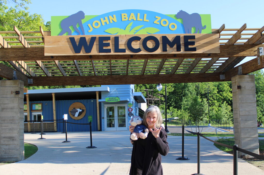 Woman standing in front of John Ball Zoo Welcome sign holding up "I love you" hand sign. She is holding a stuffed monkey.