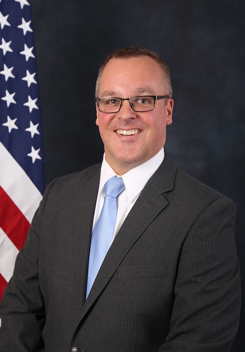 Professional photo of Matt Groesser wearing a suit with an American flag in the background