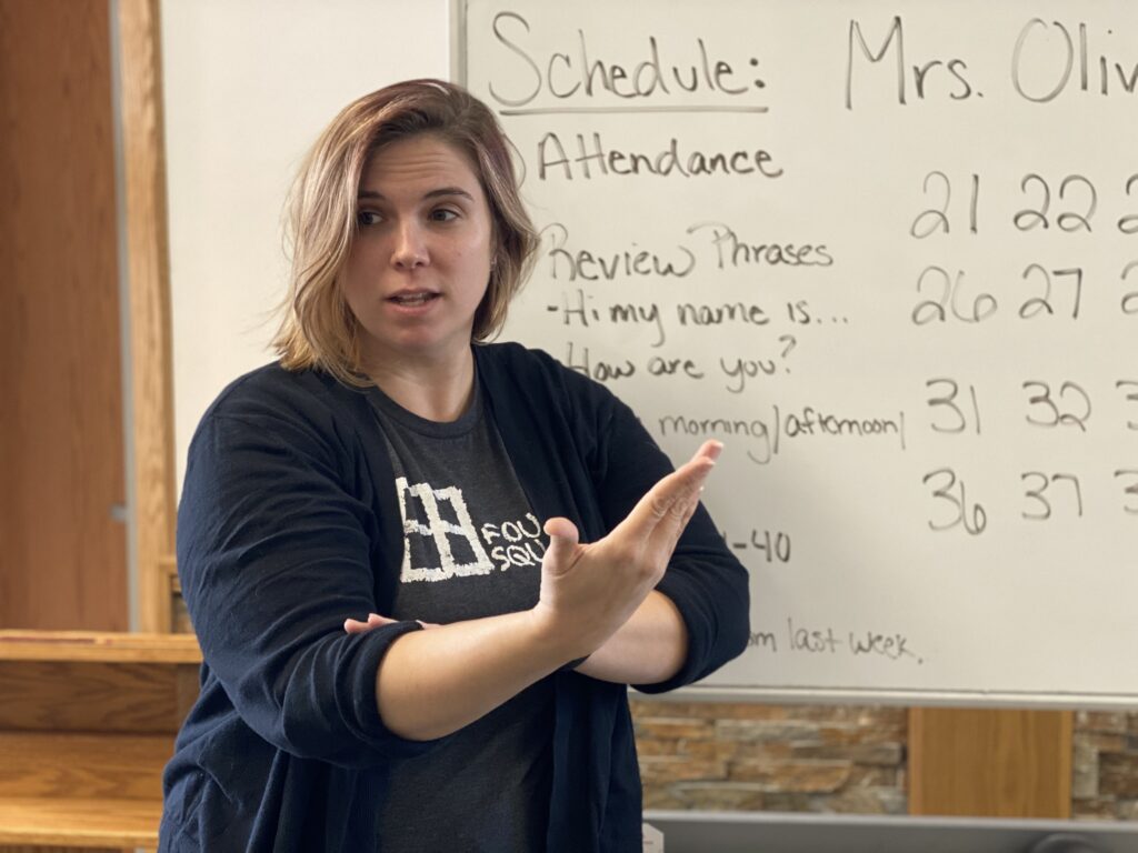 Woman with shoulder-length hair stand next to a whiteboard in a classroom. She is signing "morning"