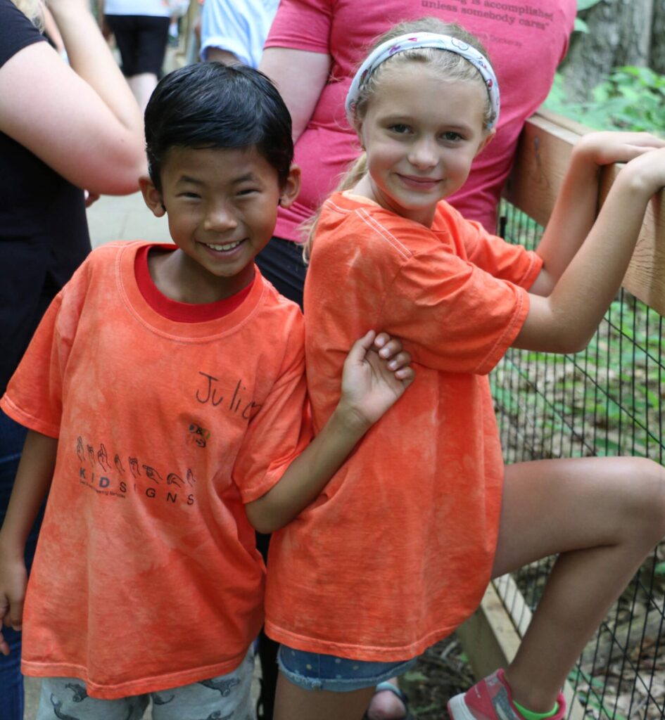A young boy and girl standing next to each other smiling. They are wearing matching orange Kids Kamp shirts
