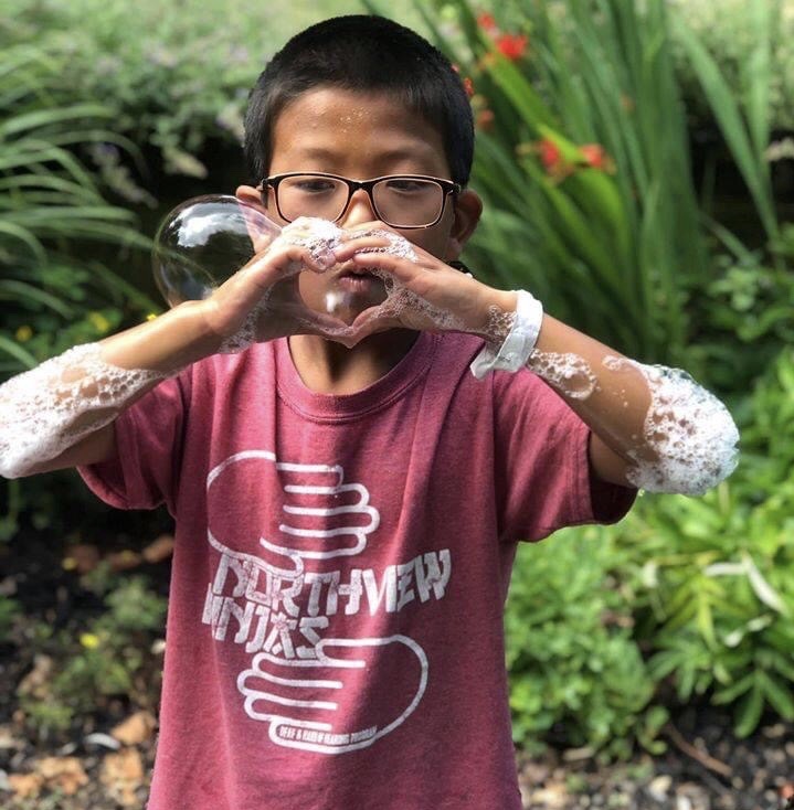 Image: Young asian boy with glasses and a red shirt blowing a bubble through his hand.