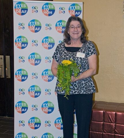 Claudia standing in front of a D&HHS banner. She is holding flowers and smiling,