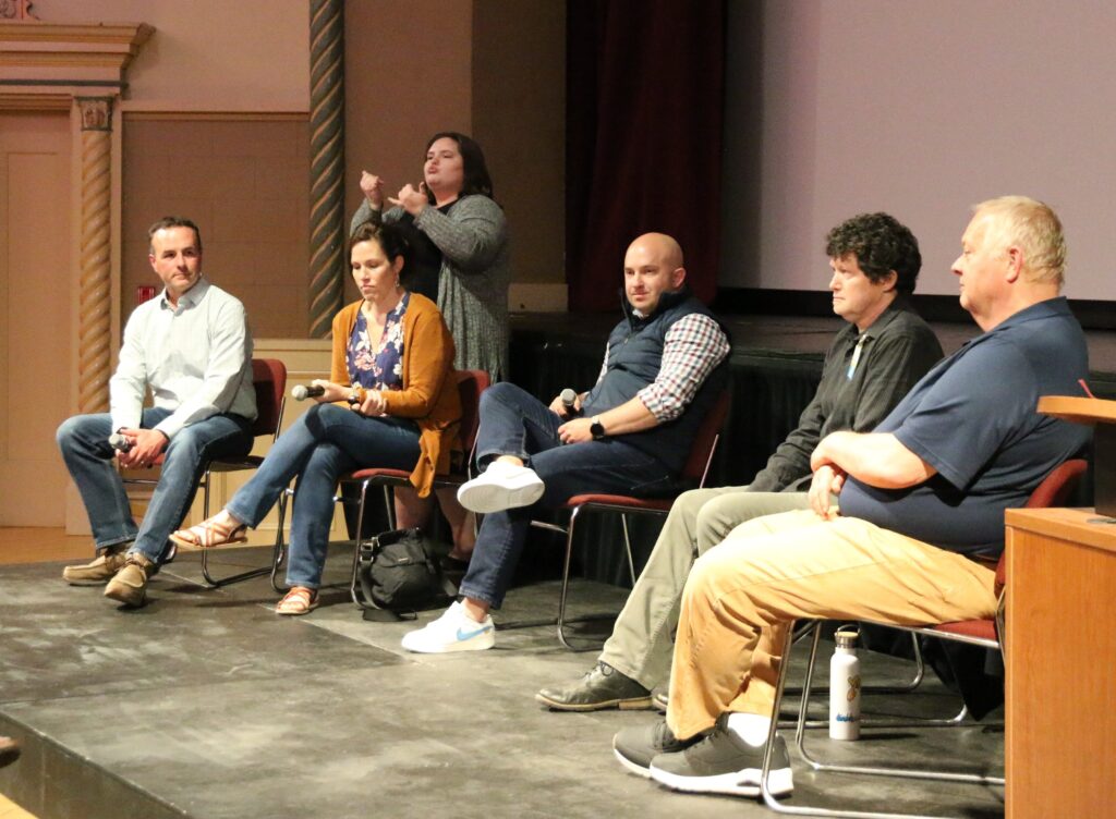 Panel of 5 people seated on a platform. An interpreter is in the left background