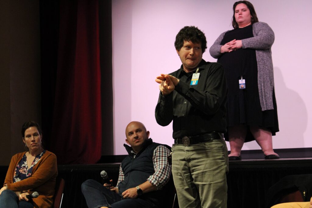 Rowan, a man wearing a black button up standing and using ASL. There are people and a theatre stage in the background.