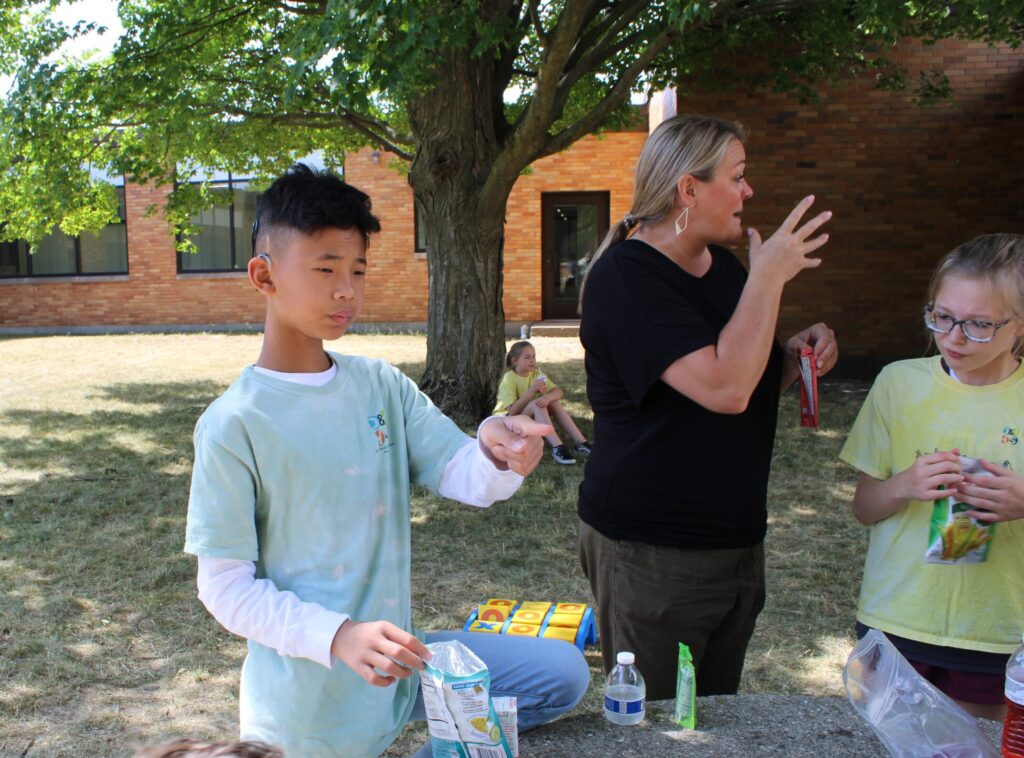 Julian standing outside in a green shirt during snack time helping. An interpreter dressed in black is standing to the right