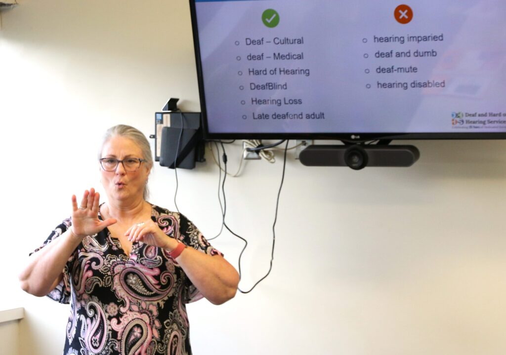 Nancy stands in front of a TV screen that shows a presentation. She is signing to the group about appropriate termonology