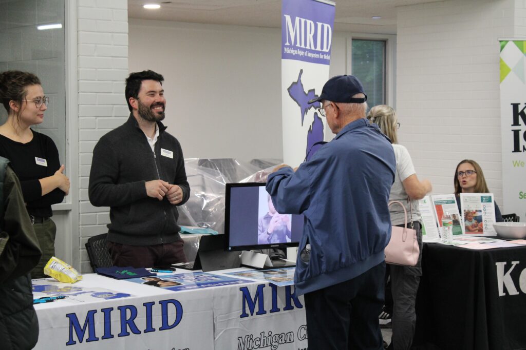 Man wearing a blue jacket and a hat is talking with two people behind a table that says "MIRID"