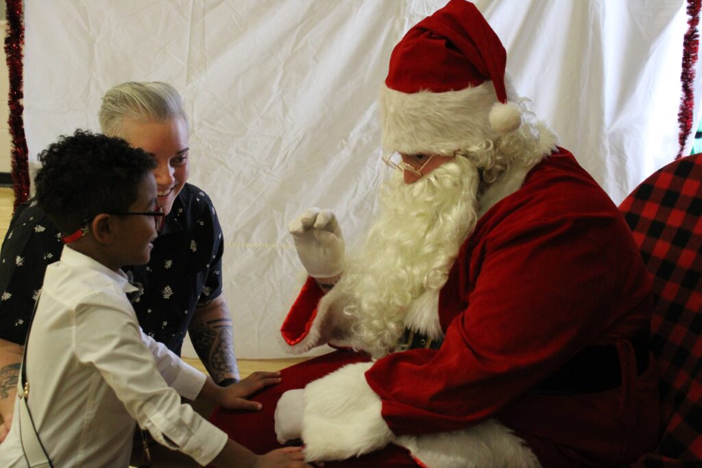A boy standing in front of Santa as he signs. A woman is in the back smiling