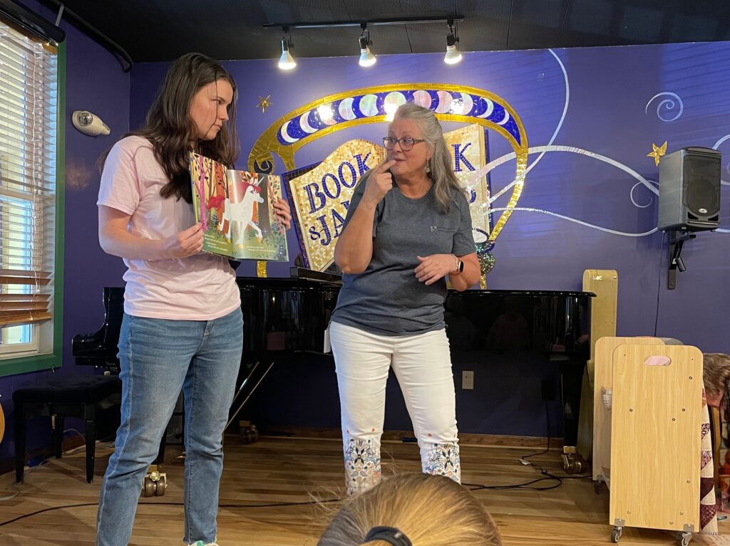 Nancy and Bri standing on a stage. Bri is holding a book while Nancy signs the story