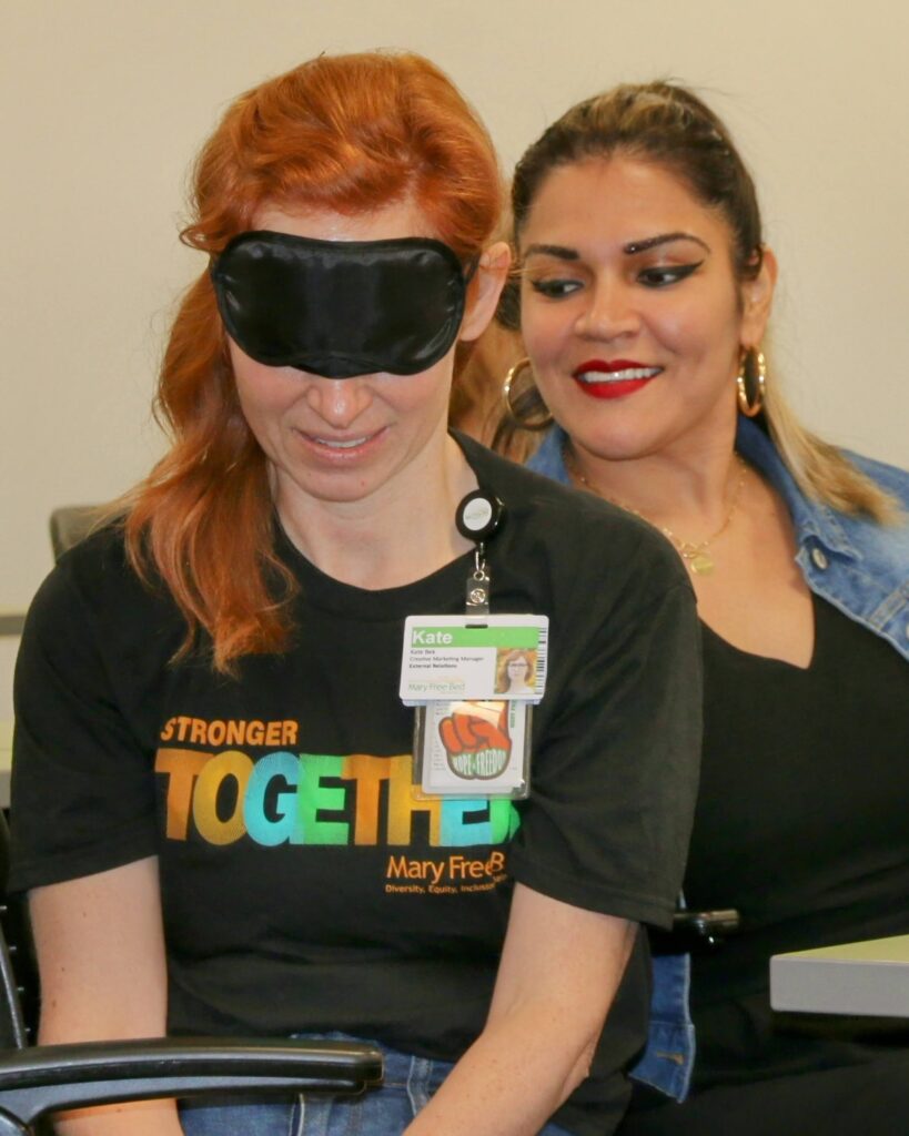 Woman with with dark hair smiles as she practices haptics on the back of another woman with red hair wearing a blindfold.