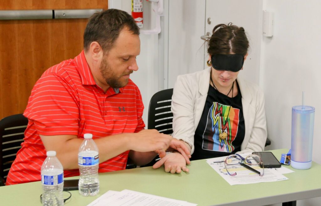 Man in a red shirt practicing print on palm with a woman who is blindfolded