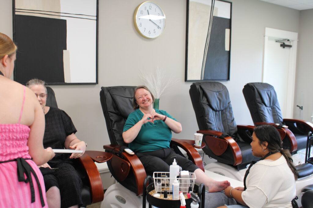 Woman sitting in pedicure chair holding up a heart hand shape ot the camera