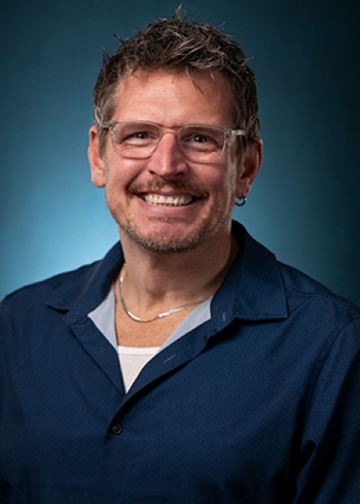 Photo of Christopher Rawlings. He is a white man wiht a goatee and light framed glasses. He is wearing a dark blue collared shirt with a light blue background.
