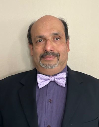 Photo of Anindya Bhattacharyya. He is an Indian man with a mustache and bald. He is wearing a purple button down shirt with a light purple bow-tie and black jacket in front of a tan background