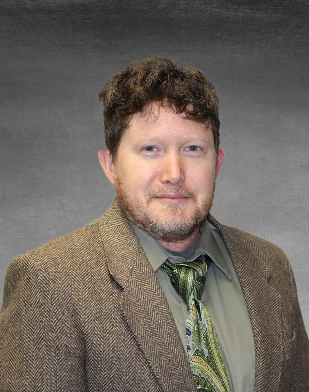 Professional headshot of Rowan smiling. He is wearing a green button up shirt with a green patterned tie and a brown blazer